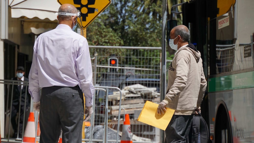 A man wearing a face mask and carrying a backpack and piece of paper walks off a bus near another man wearing PPE.