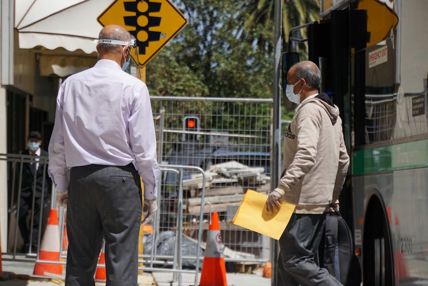 A man wearing a face mask and carrying a backpack and piece of paper walks off a bus near another man wearing PPE.