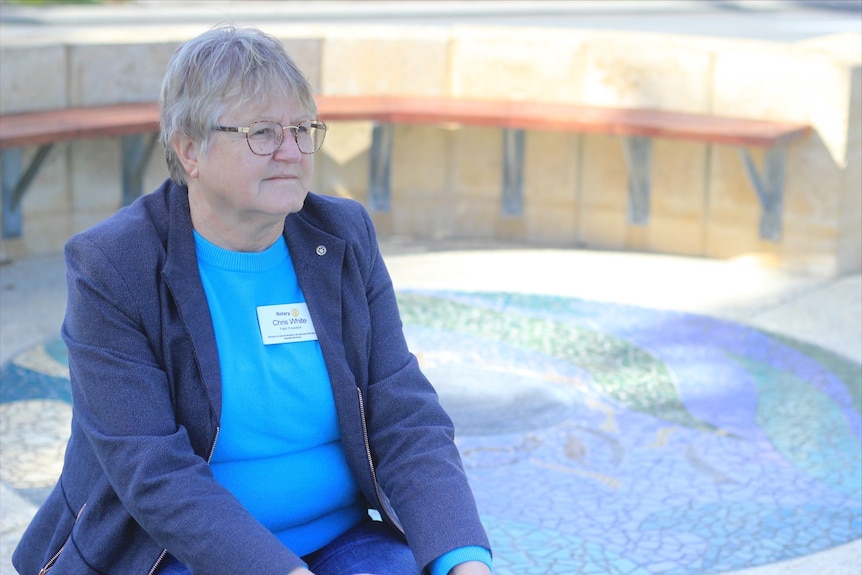 A bespectacled woman with short grey hair sitting at a memorial.