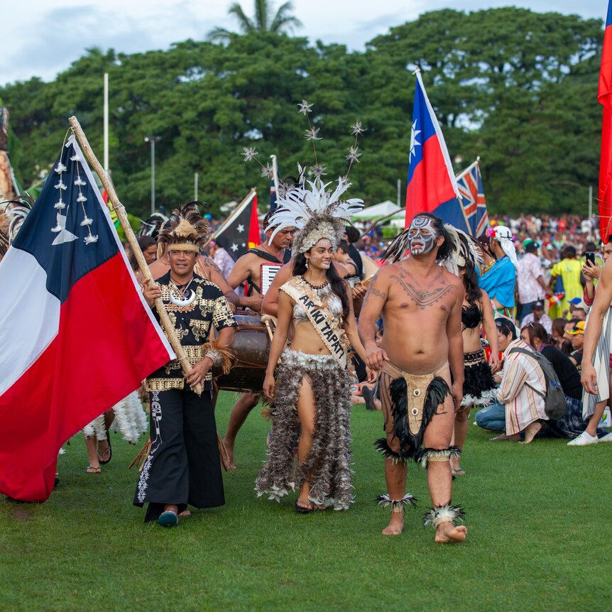 People in traditional Pacific dress march holding flags on green grass. 