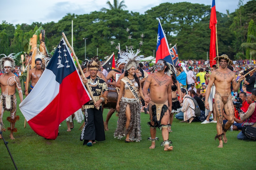 People in traditional Pacific dress march holding flags on green grass. 