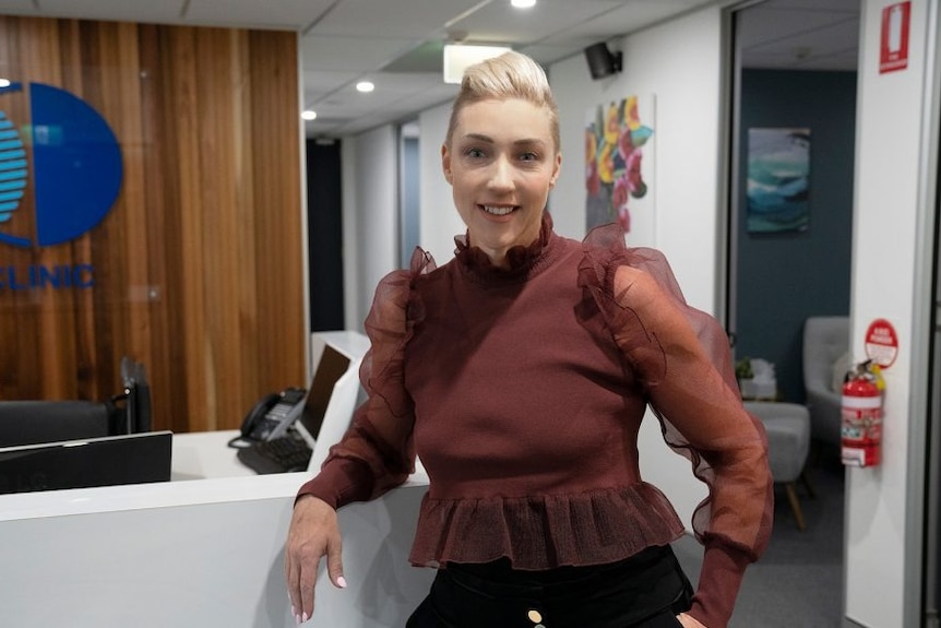 A woman with short blonde hair wearing a maroon top leans on a reception desk inside a psychology clinic.