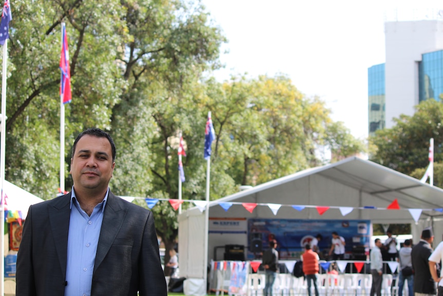 A man stands smiling against a white tent with national flags and a row of plastic chairs in the background