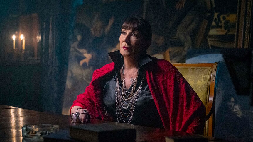 Colour still of Anjelica Huston sitting at a table in a dimly lit room in front of oil paintings in John Wick: Chapter 3.