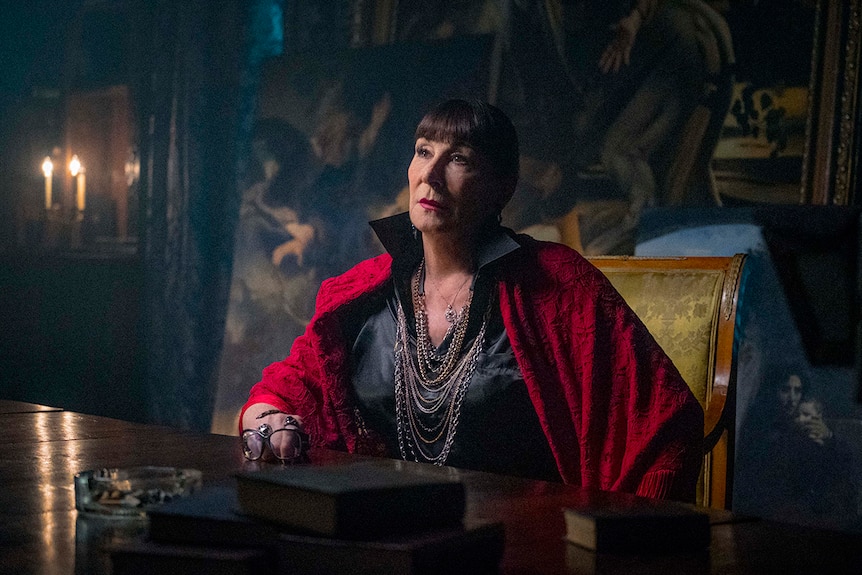 Colour still of Anjelica Huston sitting at a table in a dimly lit room in front of oil paintings in John Wick: Chapter 3.