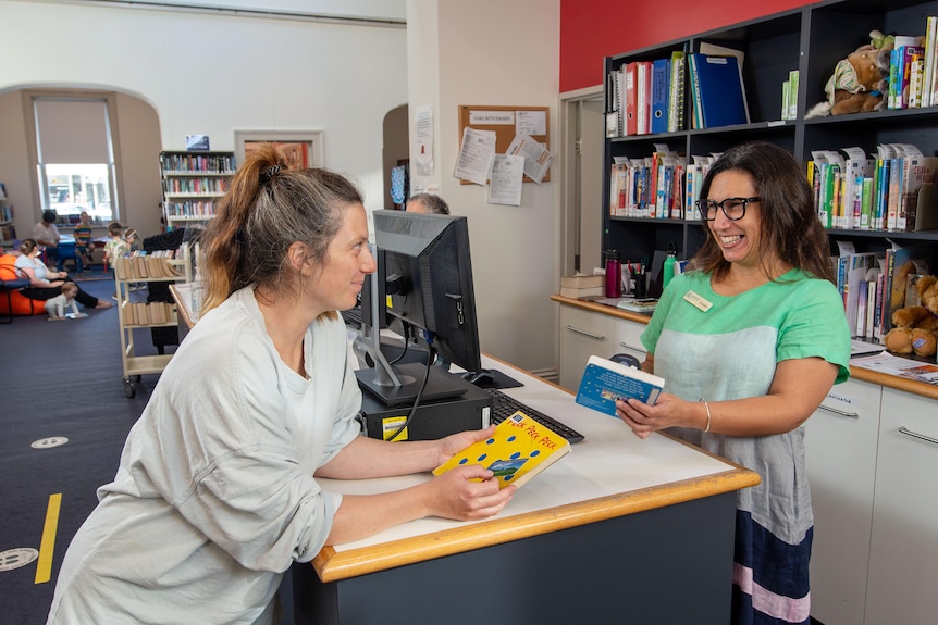 A library worker stands behind a desk chatting to and laughing with a customer borrowing books. 