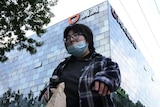 A person in a flannelette top wearing a blue face mask and glasses in front of a tall glass building on an overcast day 