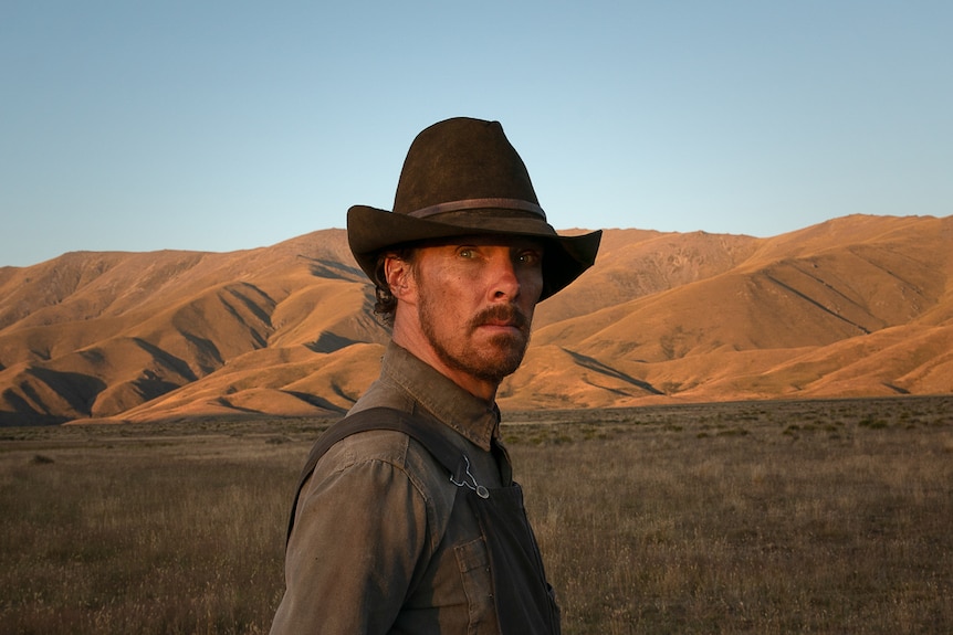 40-something white man rough-shaven dressed like rancher standing in western plain, looking at camera.