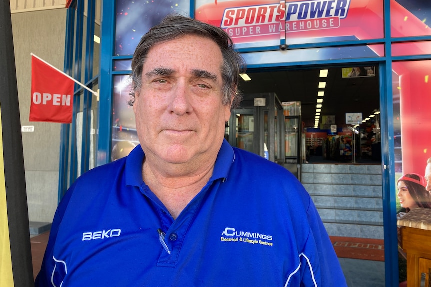 man in a blue shirt standing outside a sporting goods store with an open sign in the background