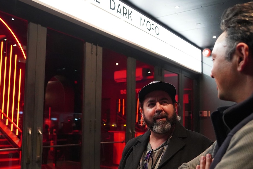 A man with a baseball cap stands under a sign which says Dark Mofo