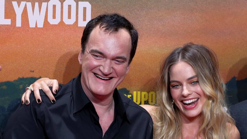 Movie director Quentin Tarantino and Aussie actress Margot Robbie pose smiling in front of a movie poster backdrop.