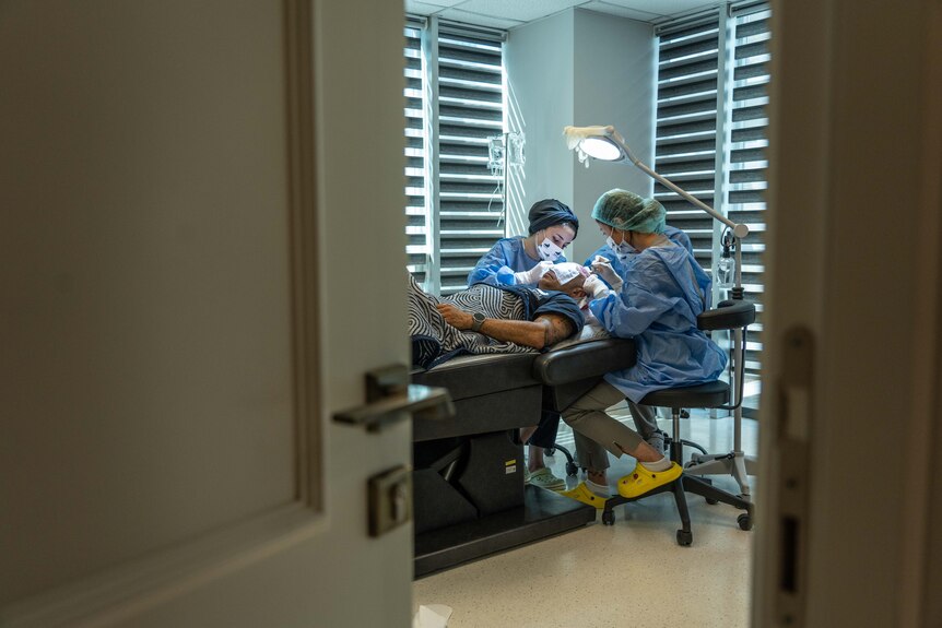 Through an open door, two nurses in surgical scrubs sit near a patient's head, using instruments on his scalp