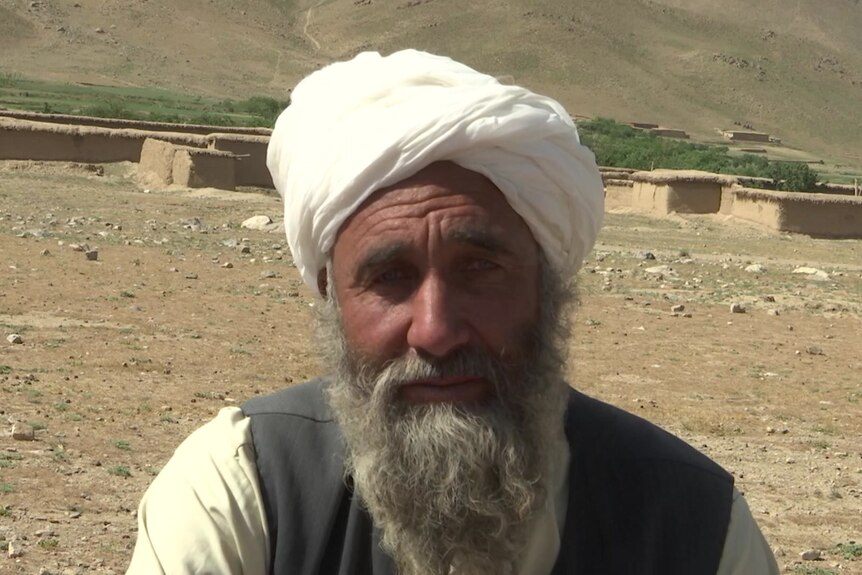 A man with a grey beard wearing a head covering sits outdoors and looks into the camera with a serious expression.