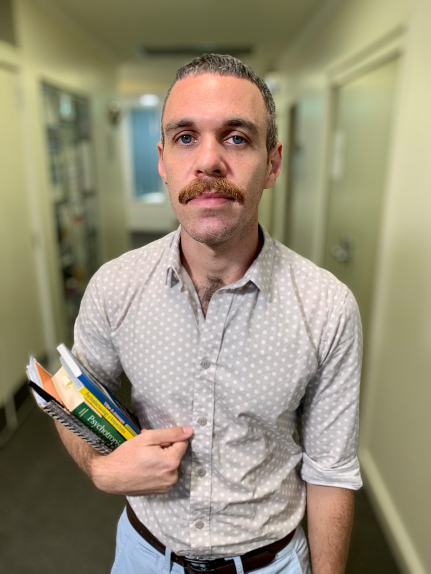 A man looking at the camera while standing in the hallway of a medical practice and holding textbooks. 