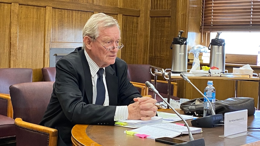 A man wearing a suit speaks into a microphone mounted on the desk he's seated at