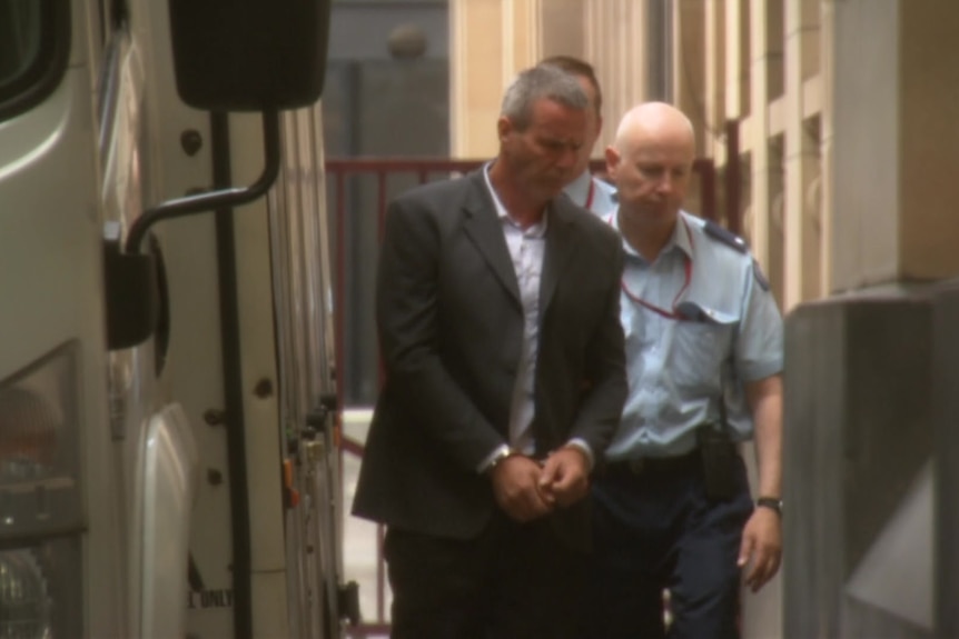 A handcuffed man in a suit is escorted towards a building by a police officer.