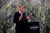 A balding, young to middle-aged man in a suit speaks behind a black podium in front of a forest-style backdrop.