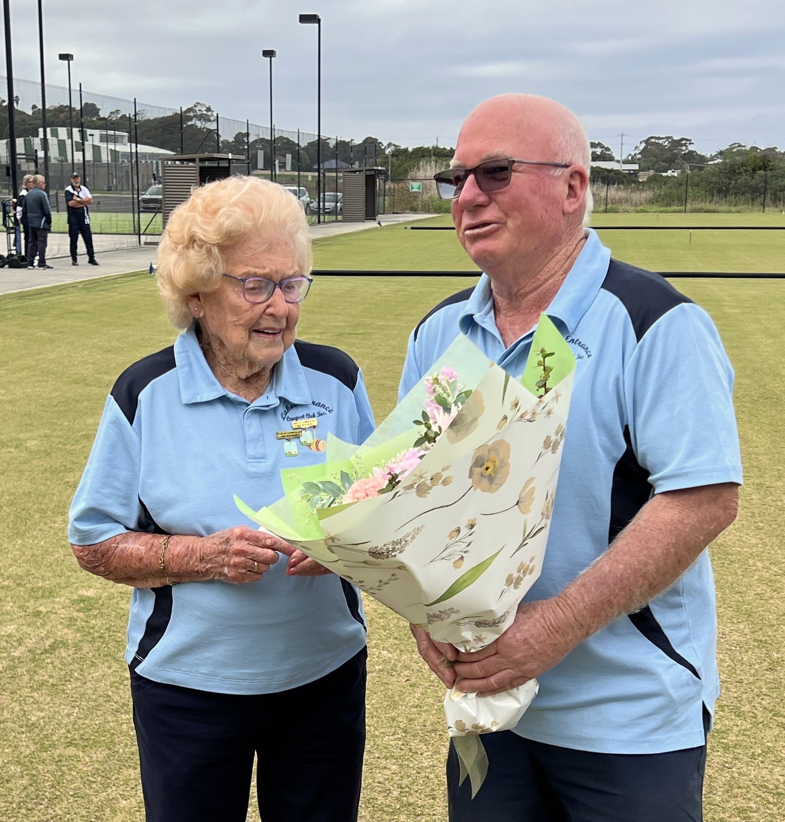 A man with white hair and glasses presents flowers to a woman with a blonde perm, on a croquet court.