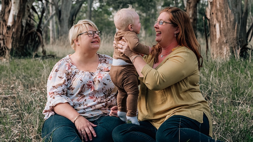 Kait lifts a laughing Arlen into the air, as she sits with Christel in the bush.