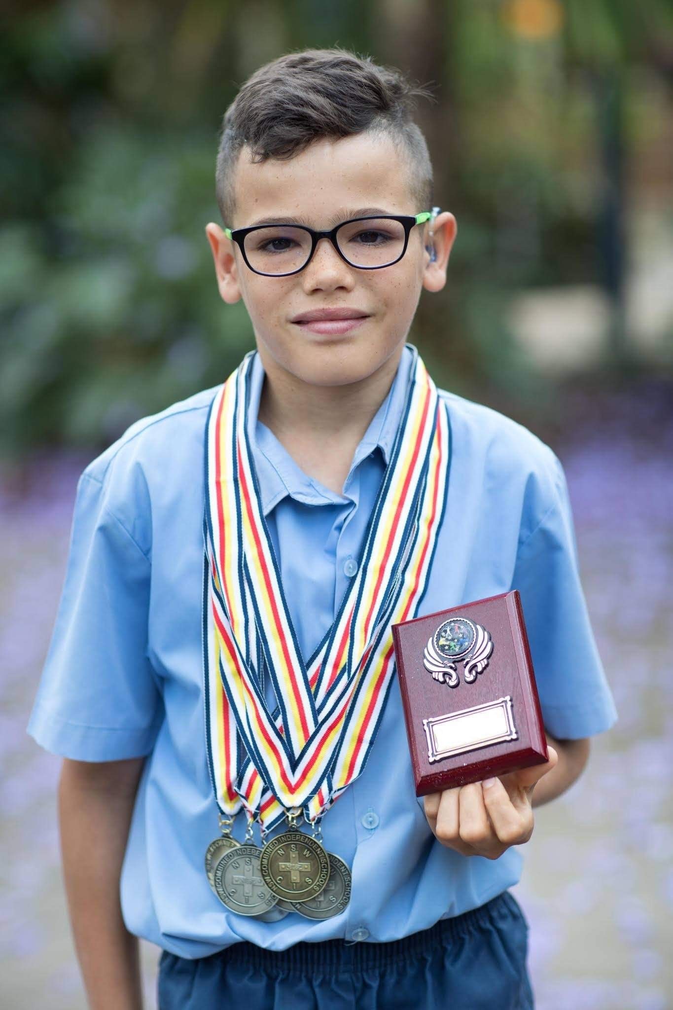 A young boy smiles as he wears multiple medals and holds a trophy.