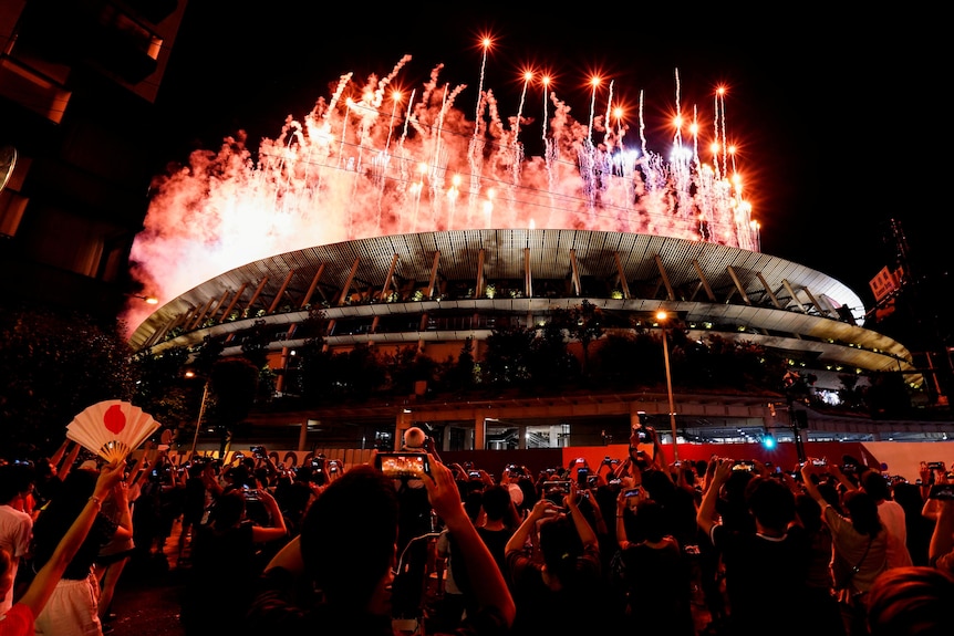 People hold their phones up to take pictures while fireworks erupt over a stadium