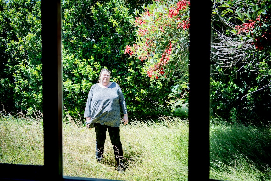 Shelly standing amid long grass, surrounded by leafy trees, in the backyard of her home.