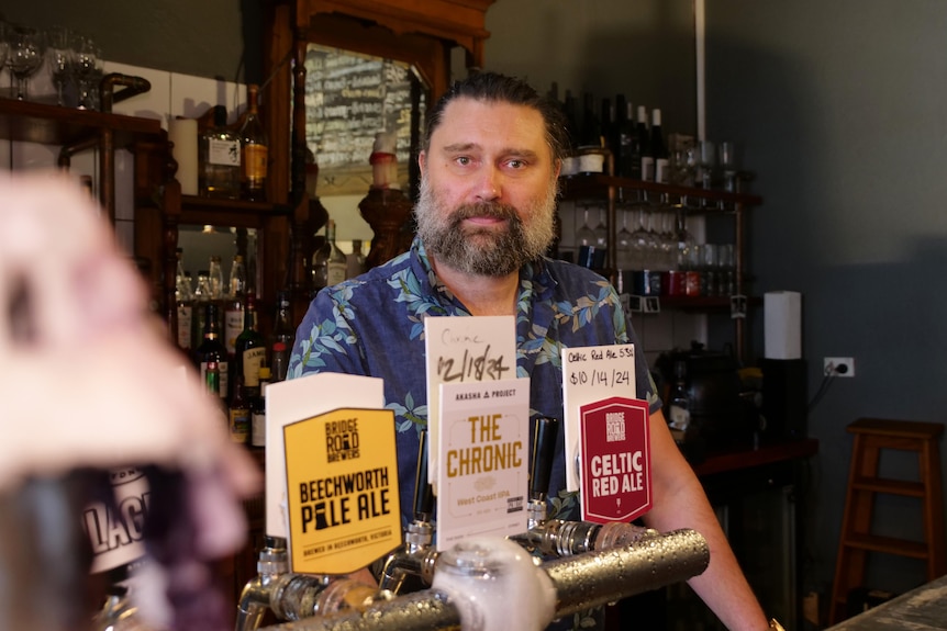 A middle-aged man with a beard stands behind the beer taps behind a bar.