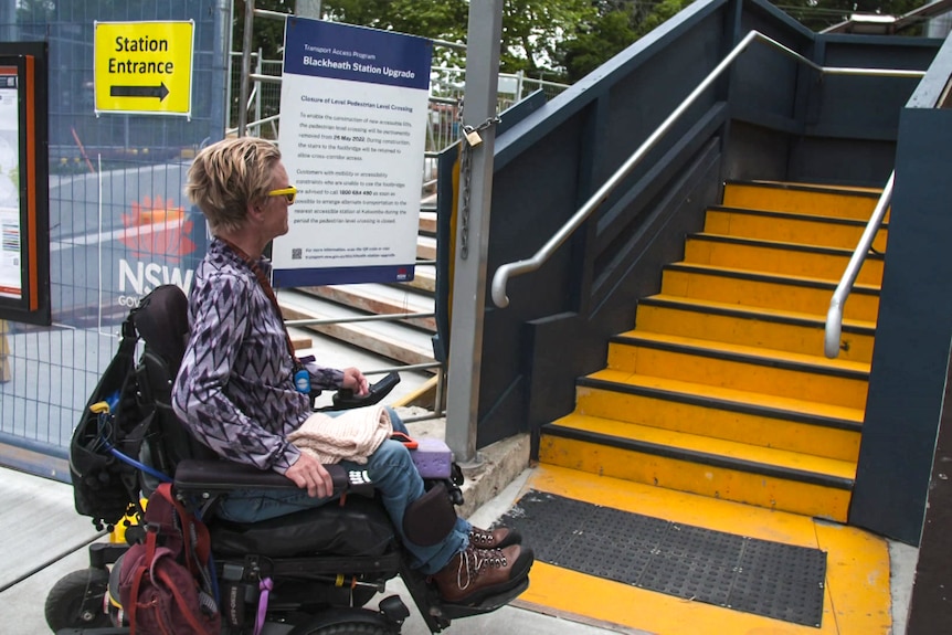 Woman in a wheelchair looks up at a set of concrete stairs at a train station