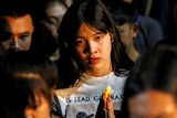 A woman stares at the camera from a crowd of people with bowed heads.