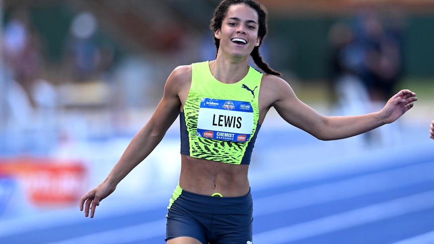 An 18-year-old female sprinter crosses the line in first place, with a smile, wearing a top and sprinting shorts.