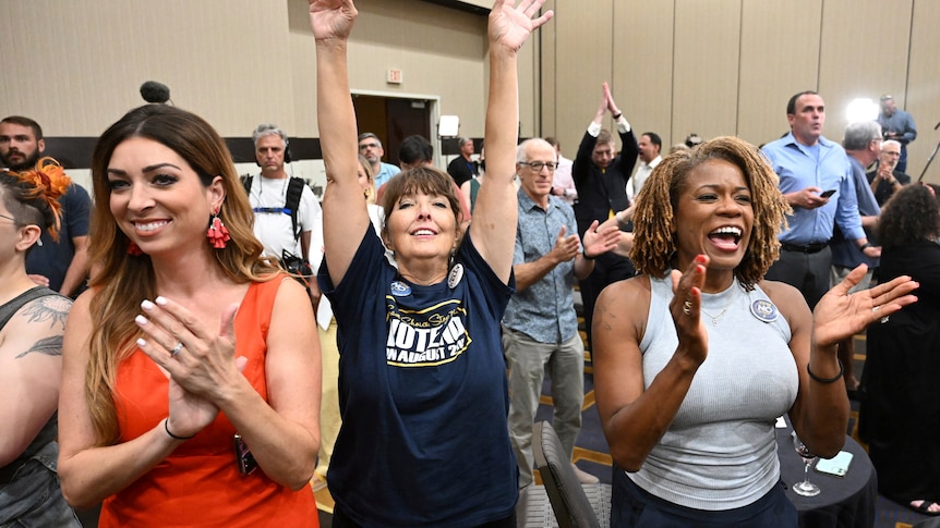 Three women at front of a conference room crowd cheer. Two clapping, one in middle with arms up