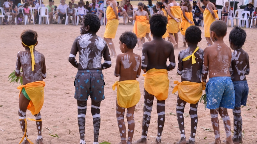 Seven Indigenous children are standing in line at a cultural festival, dancing in traditional dress.