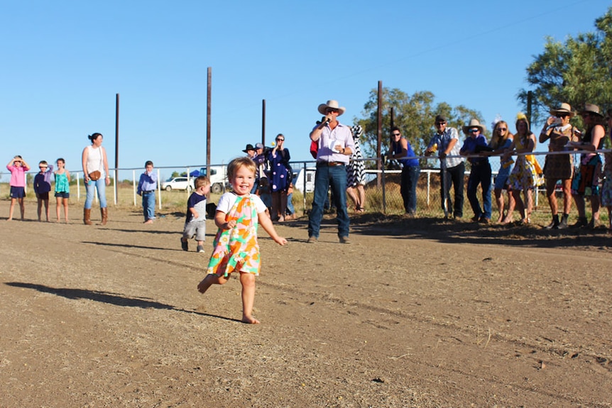 A little girl runs down a horse racing track, while a group of adults and other kids watch on.