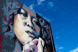 A mural of a woman's face painted on the side of a building, with blue sky in the background. 