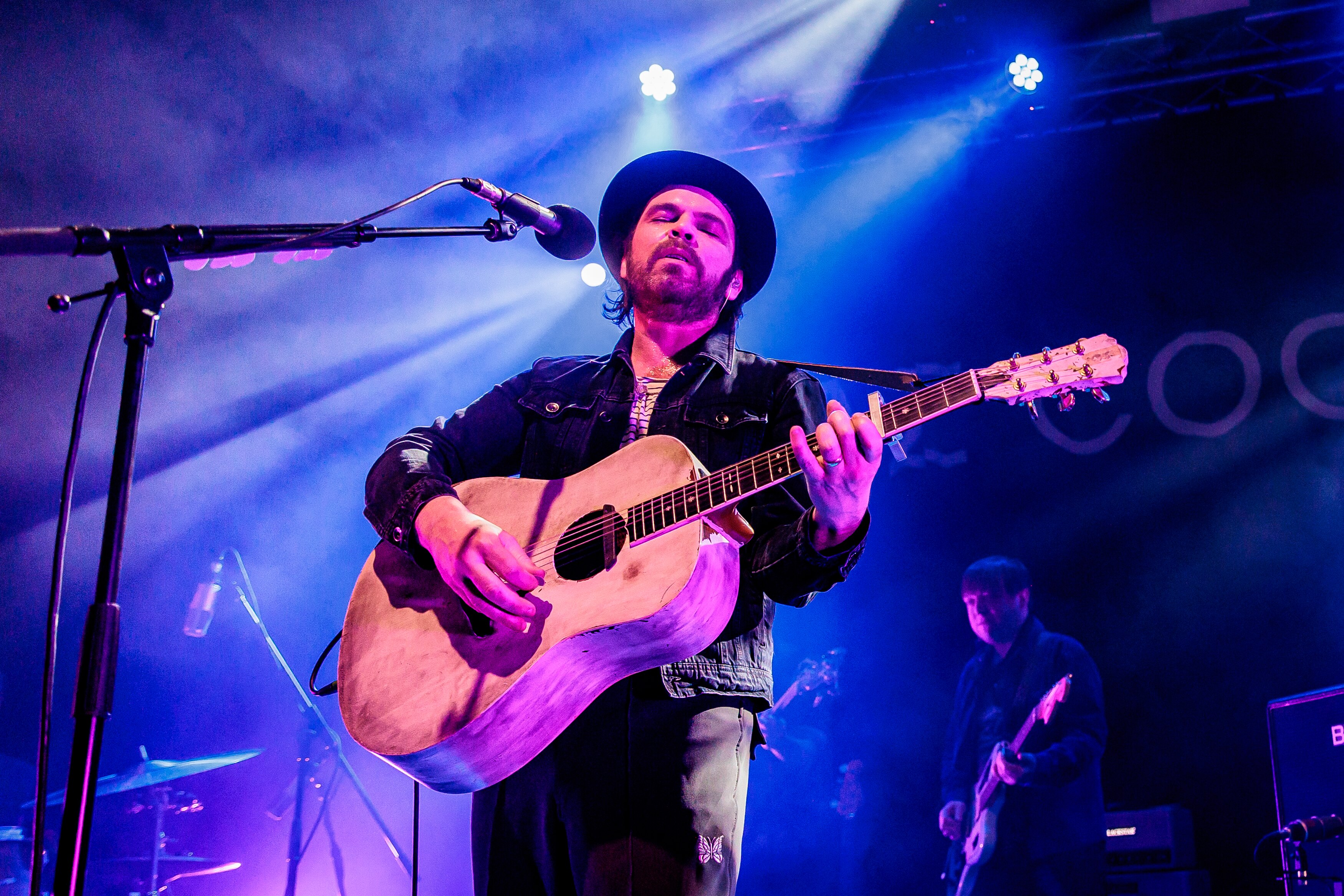 Gaz stands behind a microphone holding a guitar under blue light with his eyes closed
