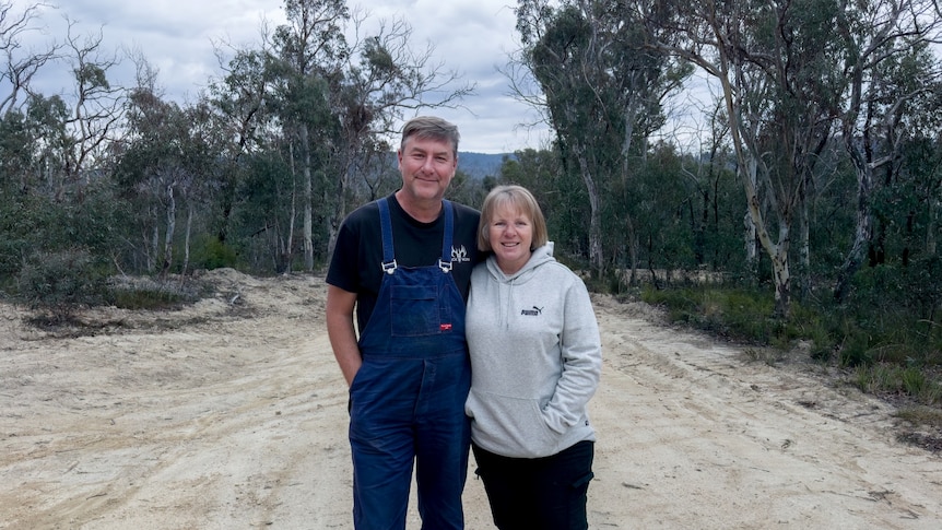 Smiling middle-aged couple stand in centre of dirt road surrounded by gum trees, man wears blue overalls, woman white hoodie.