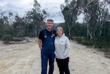 Smiling middle-aged couple stand in centre of dirt road surrounded by gum trees, man wears blue overalls, woman white hoodie.