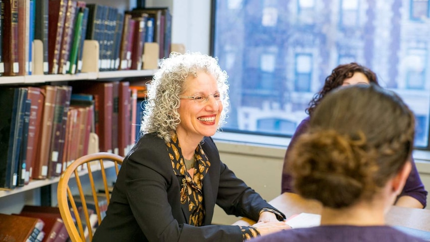 A woman sits in front of a wall of books, smiling during a group learning session.