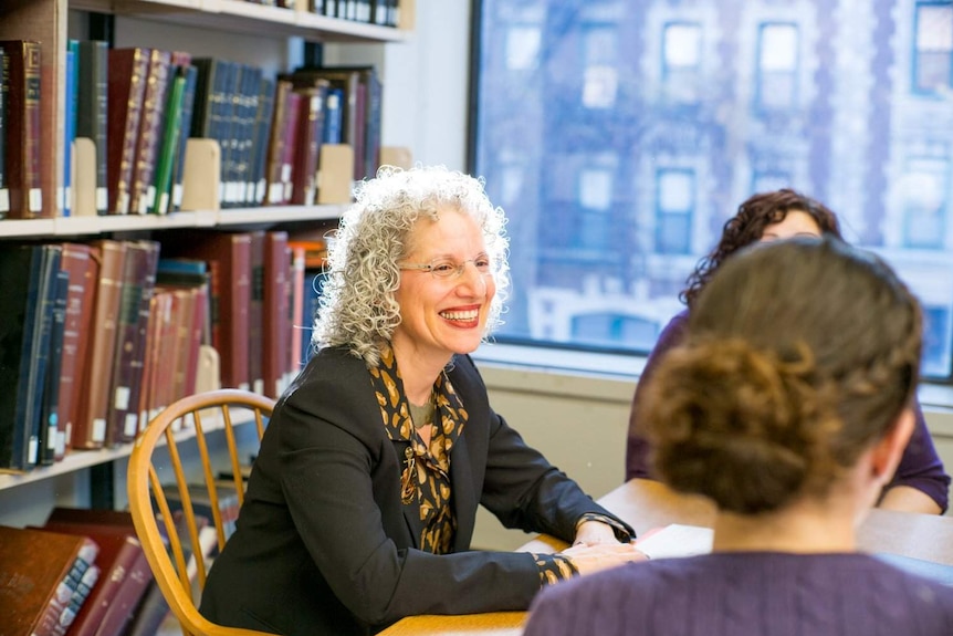 A woman sits in front of a wall of books, smiling during a group learning session.