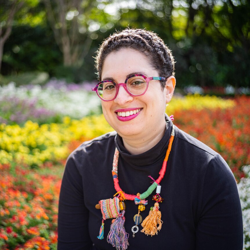Photo of a smiling woman wearing a brightly coloured necklace in front of a bed of flowers