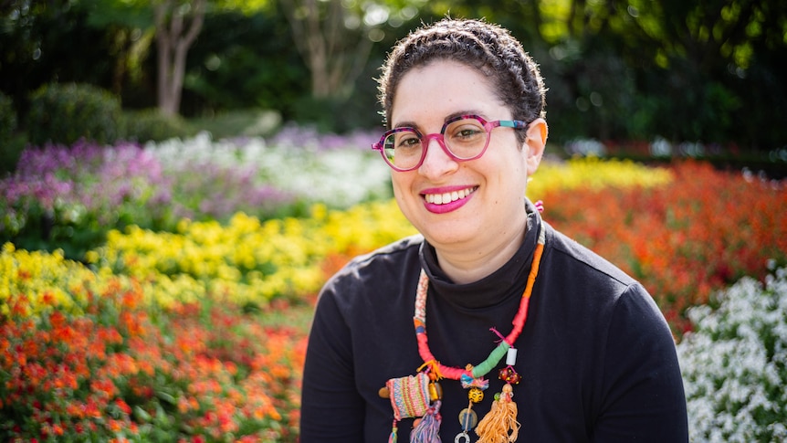 Photo of a smiling woman wearing a brightly coloured necklace in front of a bed of flowers