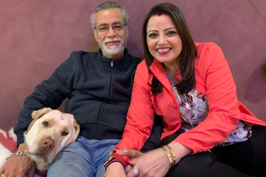 A woman and a man sit side by side in their home, a dog next to them.