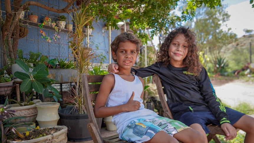 Two Indigenous boys sit on deck chair in a backyard, with one giving a thumbs up