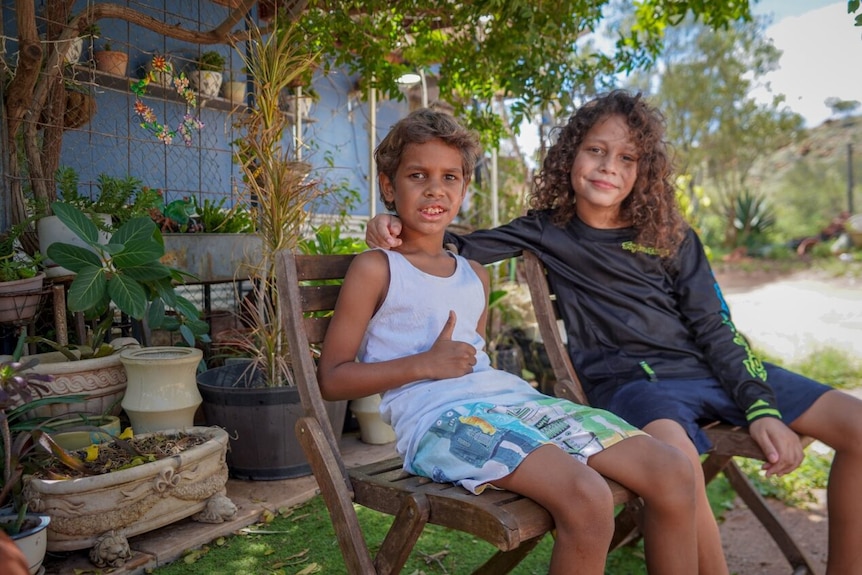 Two Indigenous boys sit on deck chair in a backyard, with one giving a thumbs up