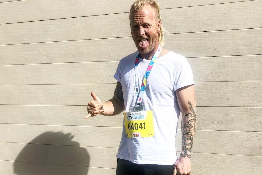 A blonde man stands with a City to Surf bib on, barefoot after completing the race.