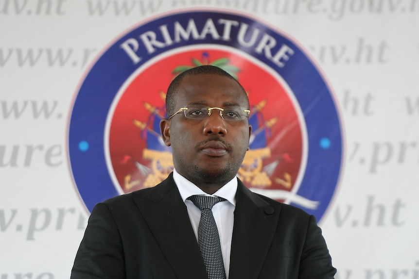 A middle-aged Haitian man in a suit with short hair and glasses stands in front of an official seal.