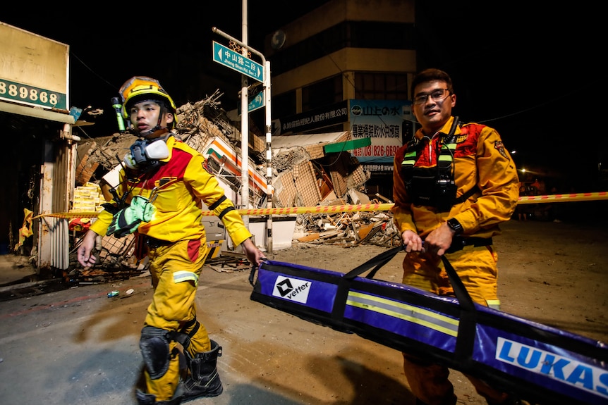 Two firefighters carrying a large tray walk in front of a collapsed building. 