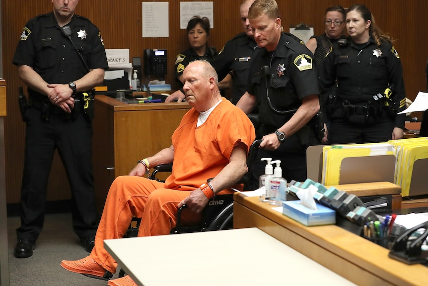 An older man in an orange prison jumpsuit, sitting on a wheelchair, is escorted through a court room by police officers.