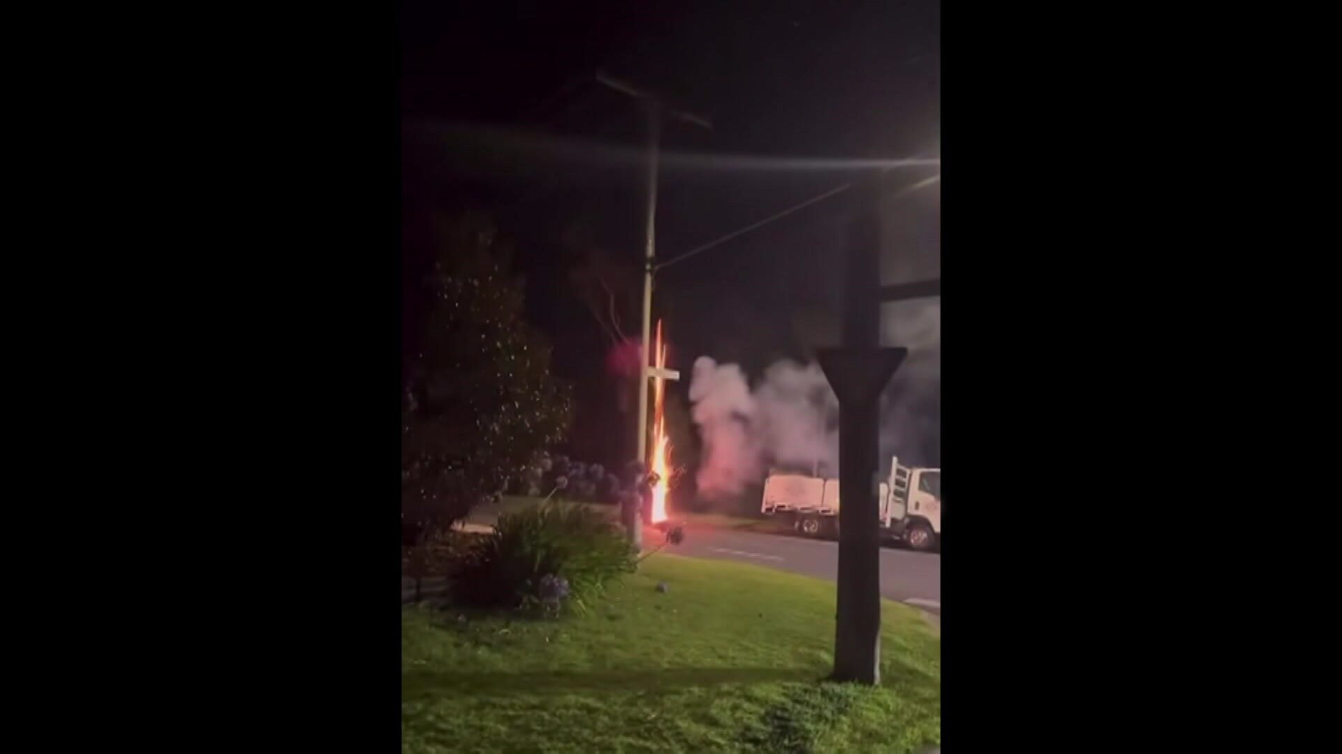 Faíscas, fogo e fumaça saem de um dispositivo em uma rua suburbana perto de um pequeno caminhão branco estacionado.