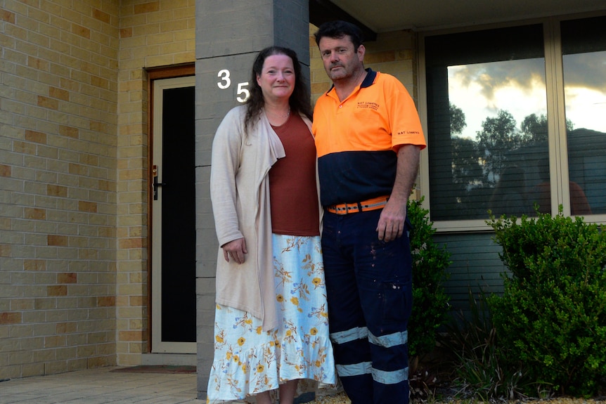 Man and woman standing outside a house. The man is wearing an orange hi-vis shirt and navy pants.